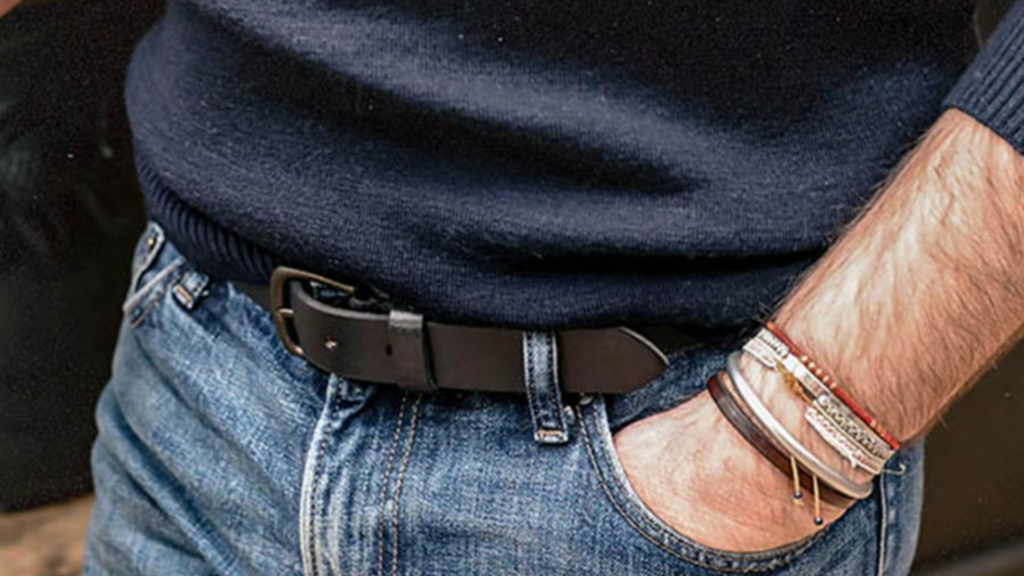 L'image montre une personne portant un pull bleu marine rentré dans un jean bleu délavé, complété par une ceinture en cuir noir avec une boucle discrète. Le bras droit est visible, avec plusieurs bracelets au poignet, mélangeant cuir et métal pour un style à la fois casual et tendance. La main est glissée dans la poche du jean, ce qui ajoute une touche décontractée à la tenue. Le look global combine des éléments simples mais bien agencés pour un style moderne et soigné.