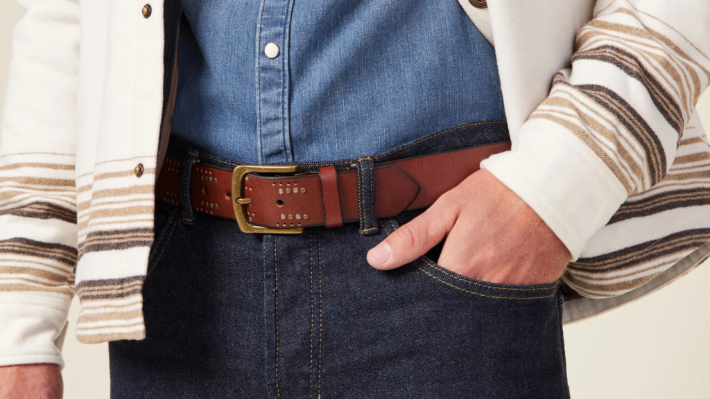 L'image montre une personne portant une chemise en denim bleu avec un jean assorti, accompagnée d'une veste blanche à rayures marron et beige. La tenue est complétée par une ceinture en cuir marron avec une boucle en laiton doré, créant un look à la fois rustique et élégant. Une main est glissée dans la poche du jean, donnant une posture décontractée. L'ensemble combine des éléments de style western avec une touche moderne et décontractée.