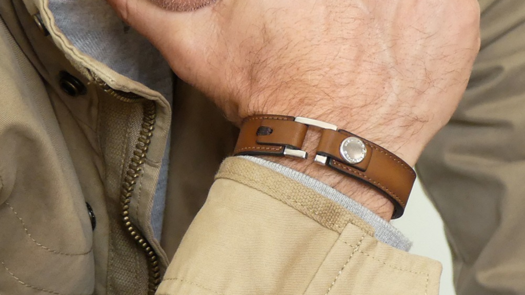 L'image montre un poignet portant un bracelet en cuir marron avec des éléments métalliques argentés. Le bracelet est élégant et simple, complétant une veste beige avec une fermeture éclair visible et un intérieur en tissu gris. La tenue évoque un style décontracté mais soigné, avec une attention portée aux détails. L'ensemble donne une impression de confort tout en restant élégant et intemporel.