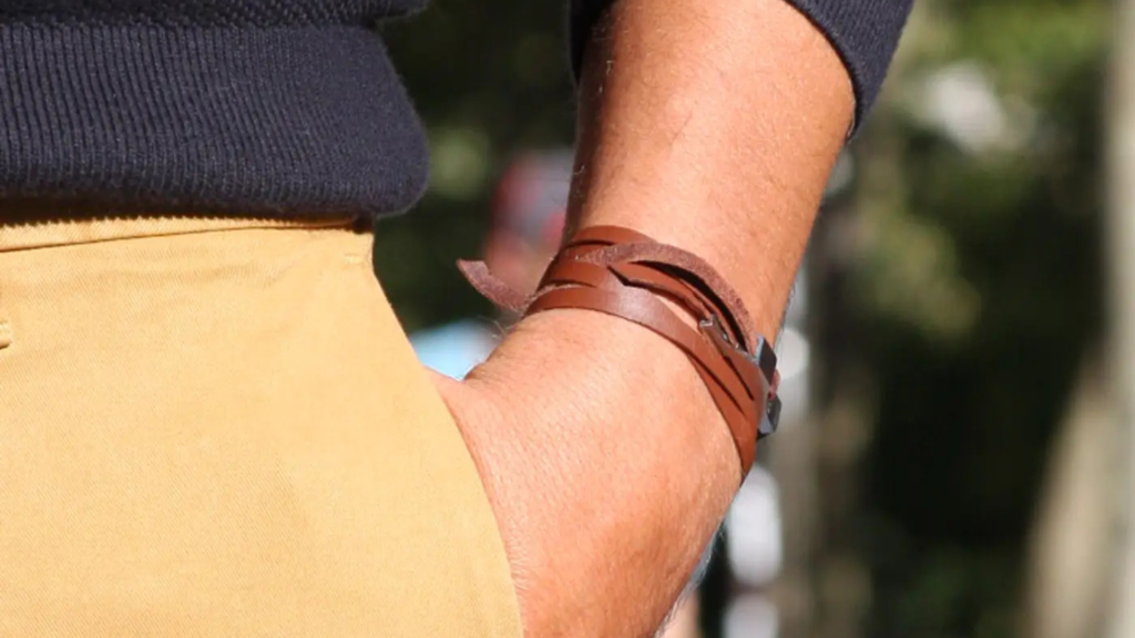 L'image montre un homme portant un bracelet en cuir marron enroulé autour de son poignet, avec une main glissée dans la poche d'un pantalon beige. Le haut du corps est vêtu d'un pull bleu marine, créant un contraste net entre les couleurs sombres du haut et les tons plus clairs du bas. Le bracelet en cuir ajoute une touche rustique et décontractée à l'ensemble, renforçant un style élégant mais simple, idéal pour une journée en plein air.