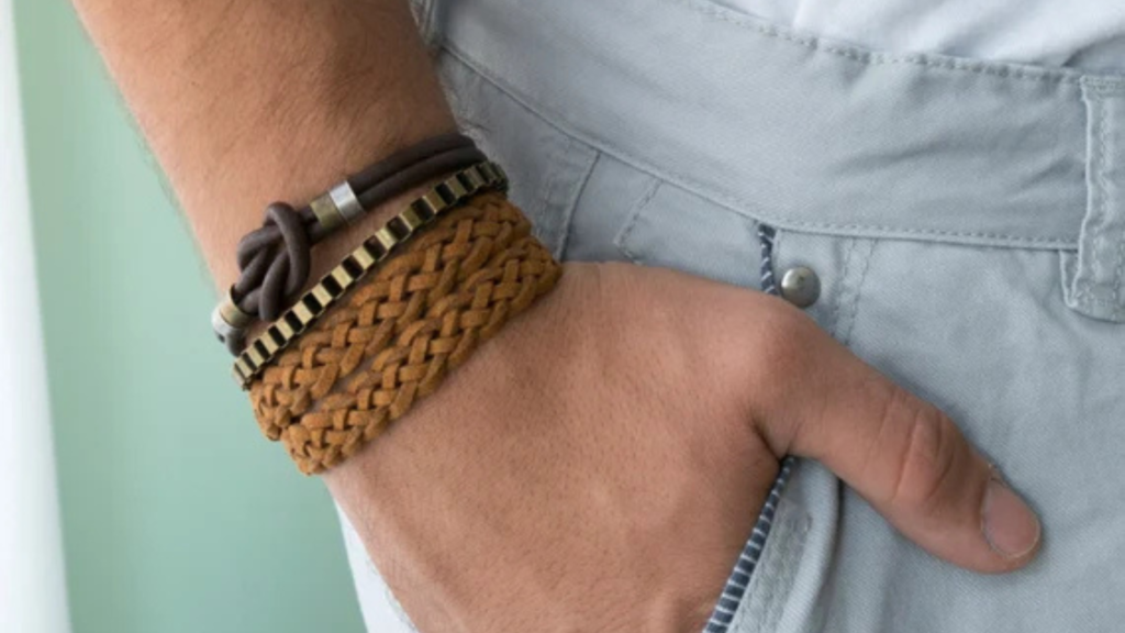 L'image montre un poignet portant plusieurs bracelets, dont un en cuir tressé marron clair et un autre en cordon brun avec des détails métalliques. La main est glissée dans la poche d'un pantalon de couleur claire, créant une apparence décontractée. Les bracelets, qui combinent des textures naturelles et des éléments métalliques, ajoutent une touche de style bohème et moderne à l'ensemble, évoquant un look à la fois simple et sophistiqué.