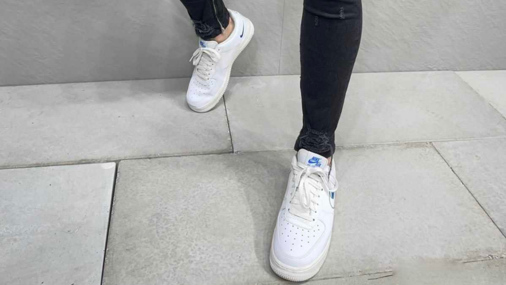 Un homme porte des baskets blanches avec des accents bleus, combinées avec un jean noir ajusté légèrement déchiré aux chevilles. L'image se concentre sur ses jambes et ses chaussures, capturant un style urbain décontracté. Les baskets, probablement d'une marque sportive populaire, ajoutent une touche classique et intemporelle au look, tandis que le jean noir usé contribue à une esthétique moderne et tendance. L'arrière-plan de sol en dalles grises renforce l'ambiance minimaliste de l'image.