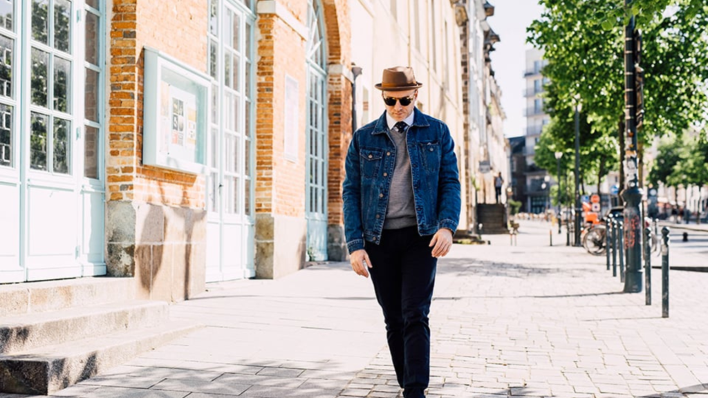 L'image montre un homme marchant sur un trottoir pavé, vêtu d'une veste en denim bleu, d'un pull gris et d'un pantalon sombre. Il porte un chapeau marron et des lunettes de soleil, ajoutant une touche élégante à son style décontracté. L'arrière-plan est constitué de bâtiments en briques avec de grandes fenêtres et une rue bordée d'arbres, évoquant une ambiance de ville calme et ensoleillée. Sa tenue, un mélange de casual et de chic, s'adapte parfaitement à l'atmosphère urbaine et décontractée de la scène.