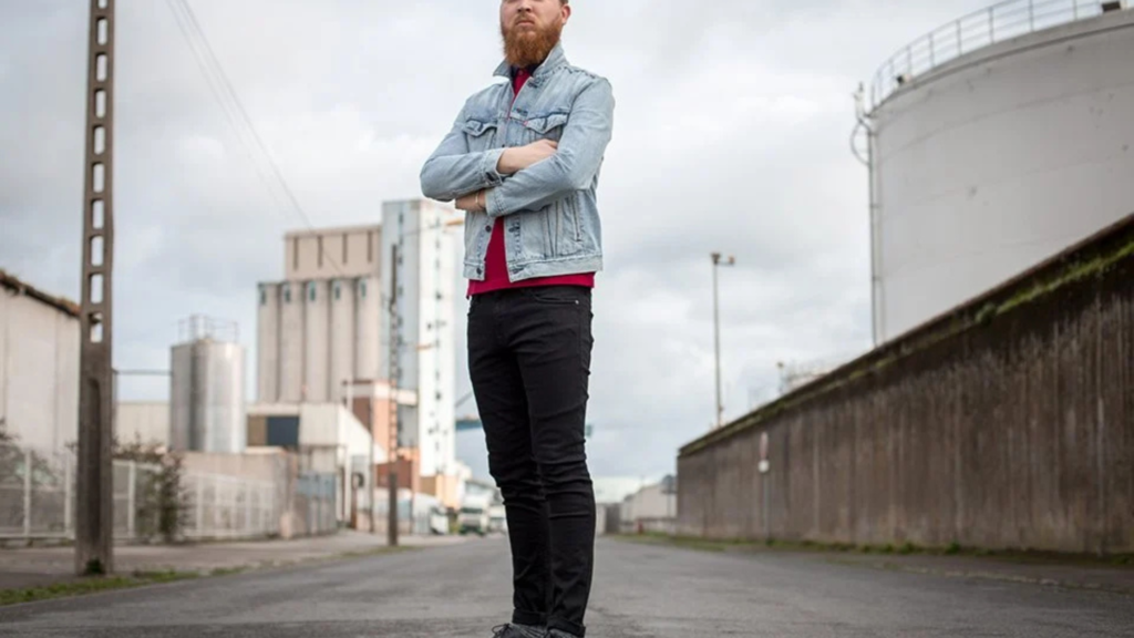 L'image montre un homme debout au milieu d'une rue industrielle, les bras croisés. Il porte une veste en jean bleu clair par-dessus un t-shirt rouge et un pantalon noir. Il est entouré de bâtiments industriels, de réservoirs et de structures en métal, créant une atmosphère brute et urbaine. Le ciel est nuageux, ajoutant à l'ambiance austère de l'environnement. Son style décontracté, avec la veste en jean et le t-shirt coloré, contraste avec l'arrière-plan industriel et sombre.