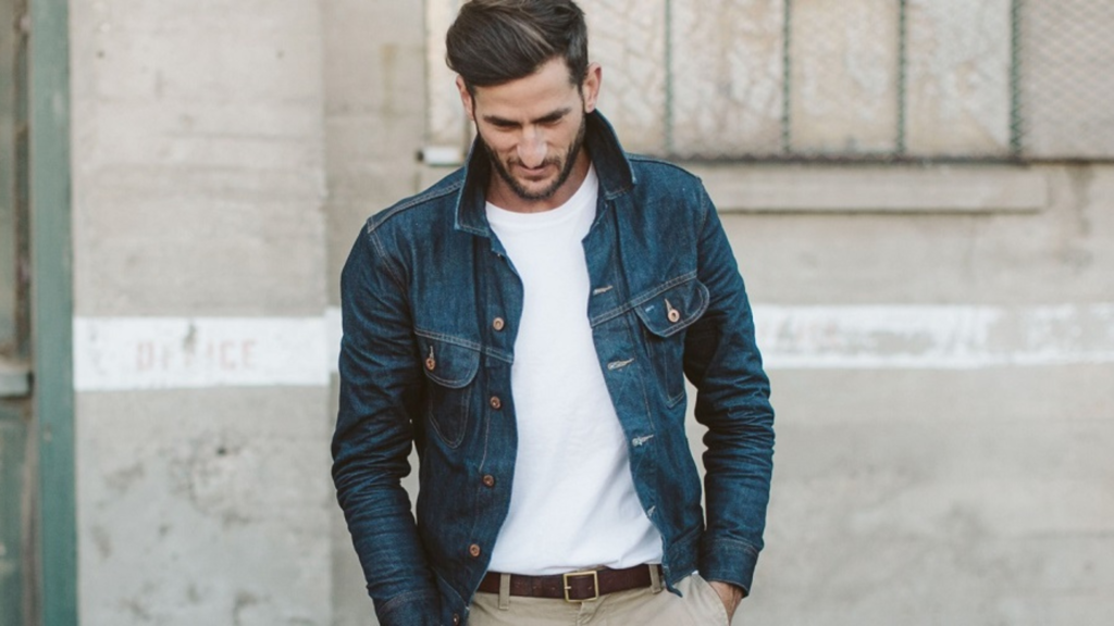 L'image montre un homme vêtu d'une veste en denim bleu foncé par-dessus un t-shirt blanc simple, avec un pantalon beige et une ceinture marron. Il adopte une posture décontractée, les mains dans les poches, tandis qu'il marche ou se tient devant un mur en béton vieilli. Son look allie des éléments classiques et intemporels, avec la veste en jean et le t-shirt blanc offrant une combinaison sobre mais élégante. L'arrière-plan industriel met en valeur le contraste entre la simplicité de la tenue et l'environnement brut, soulignant un style casual et soigné.