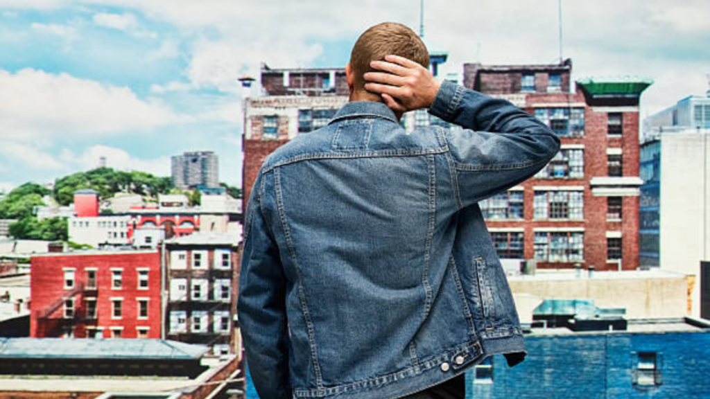 L'image montre un homme de dos portant une veste en jean bleu clair, debout face à une vue urbaine composée de bâtiments en briques rouges et de structures industrielles. Il a une main posée sur l'arrière de sa tête, ce qui donne une impression de réflexion ou de contemplation. Le ciel bleu avec des nuages blancs ajoute une atmosphère lumineuse à la scène. L'homme, dans son style décontracté, semble observer la ville, évoquant une sensation de calme ou de méditation dans un cadre urbain animé.