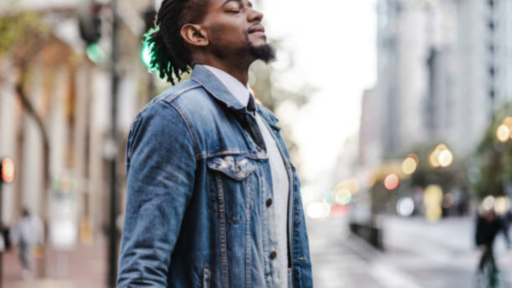 L'image montre un homme debout dans une rue urbaine, portant une veste en denim par-dessus une chemise et un pull léger. Il a les yeux fermés et semble profiter du moment, avec une expression sereine. L'arrière-plan est flou, montrant un environnement urbain avec des lumières de la ville et des passants en arrière-plan, créant une atmosphère paisible au milieu de la ville. Son look décontracté et moderne, combiné à l'environnement urbain, évoque un moment de tranquillité dans un cadre actif.